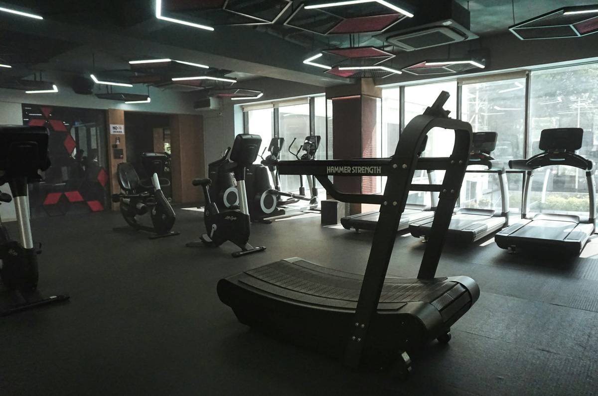 A sleek, modern fitness center featuring high-end cardio machines and strength equipment under distinctive geometric LED lighting. The space combines a professional industrial aesthetic with floor-to-ceiling windows for a bright, motivating atmosphere.