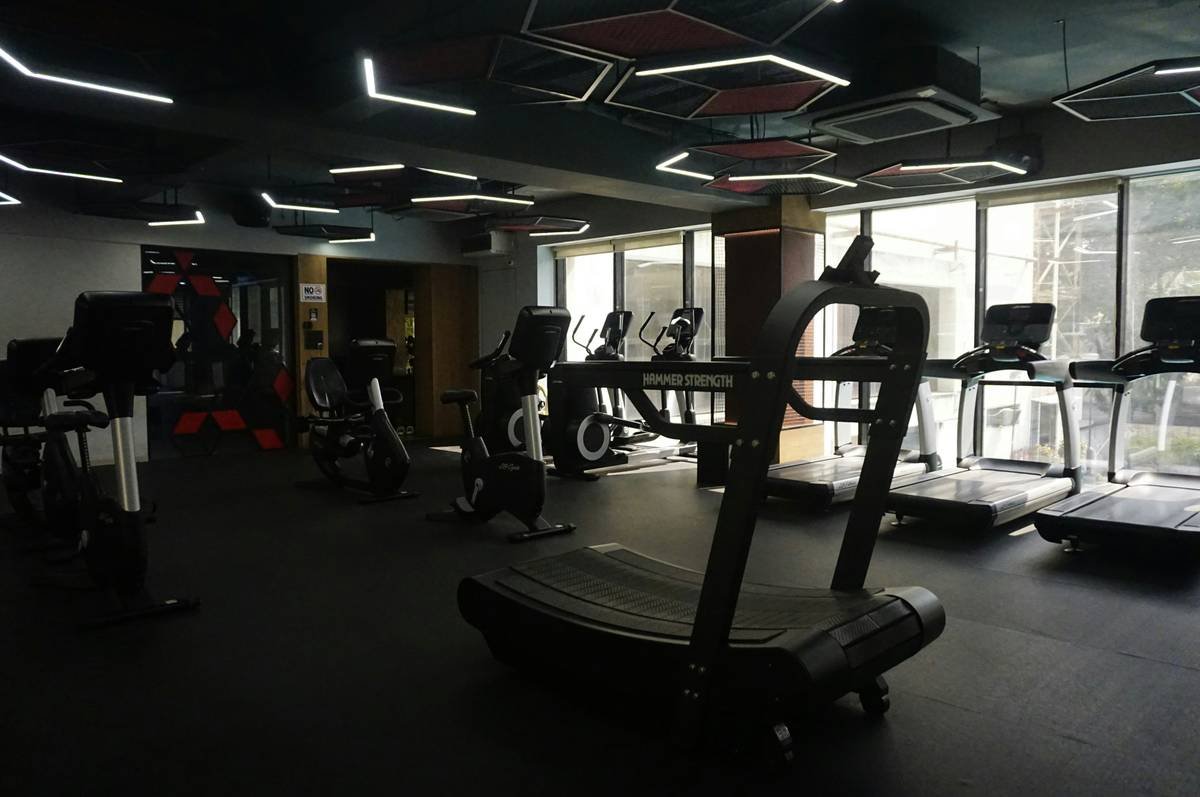 A sleek, modern fitness center featuring high-end cardio machines and strength equipment under distinctive geometric LED lighting. The space combines a professional industrial aesthetic with floor-to-ceiling windows for a bright, motivating atmosphere.