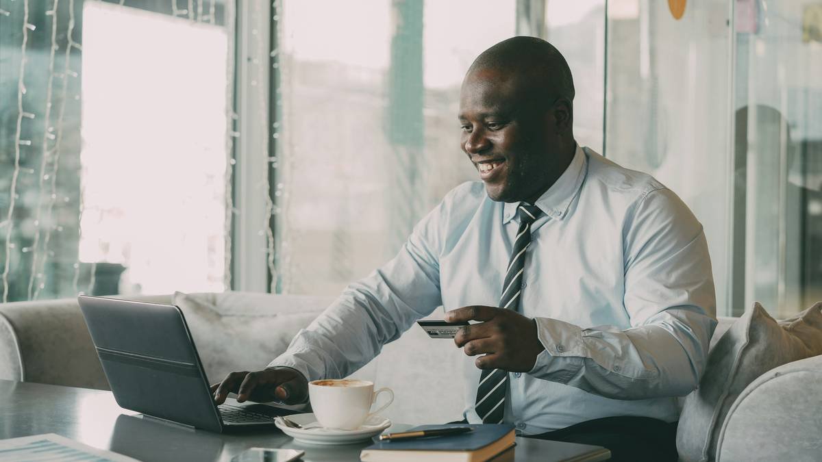 Optimistic African American businessman in formal clothes paying online bill using his credit card and laptop in fashionable cafe during lunch breaak. He smiles happily and raises his clenched fist.